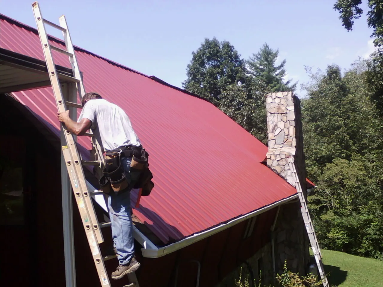 Roofer on a ladder beside a red metal roof and stone chimney — Soffit & Fascia Repair by Roofing Brothers in Berryton